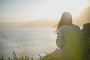 womans-back-sitting-on-cliff-overlooking-the-ocean